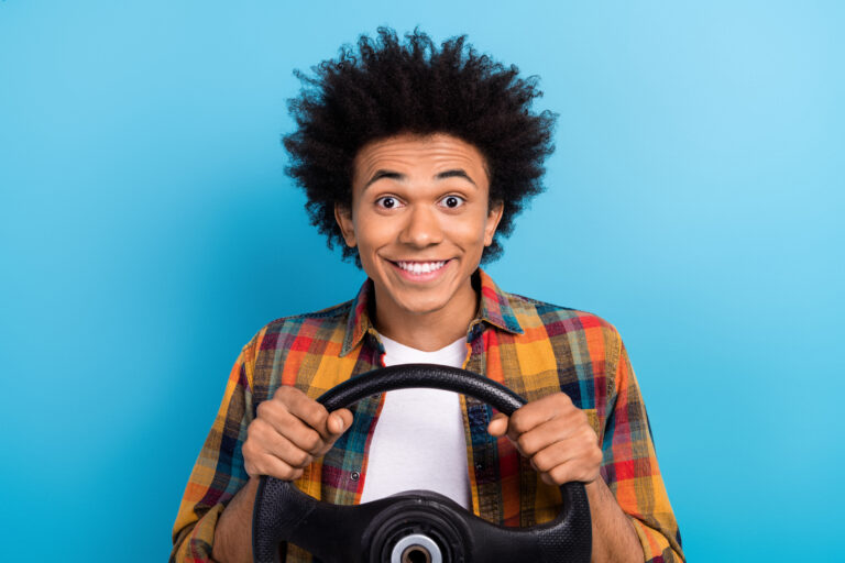 Portrait of satisfied positive guy with perming coiffure dressed plaid shirt hands hold steering wheel isolated on blue color background.